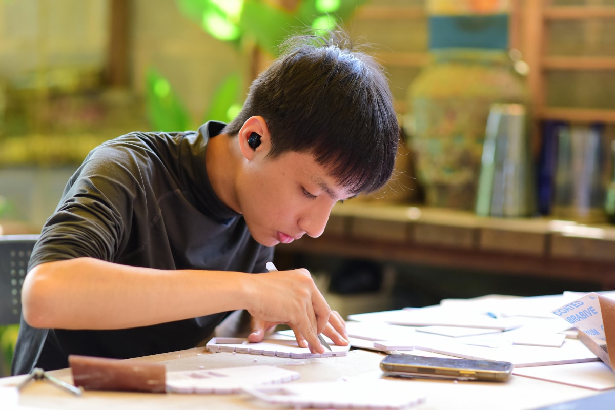 A teenage boy concentrates as he cuts pieces of foam board for a model at a busy worktable